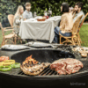 Foto da Churrasqueira a Lenha Premium Hergom com Base Alta, carne e peixe assando, pessoas ao fundo.