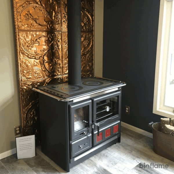 Fogão a Lenha em Ferro Fundido - Nordica Extraflame Rosa XXL com placa dourada atrás, em cozinha moderna.